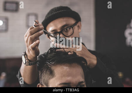 Close up ritratto professionale di barbiere rendendo il taglio di capelli del suo uomo attraente costumista con forbici e pettine barbiere vintage Foto Stock