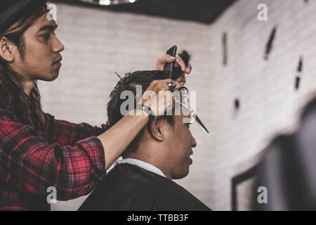 Ritratto di professional uomo asiatico con capelli lunghi marrone lavoro come barbiere rifilatura il suo cliente con forbici e pettine barbiere Foto Stock