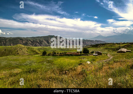La strada che conduce al borgo di montagna che si affaccia sulla Gegham creste e la sagoma del monte Ararat contro il blu del cielo coperto di nuvole Foto Stock
