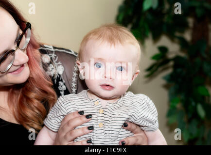 Red-headed Baby Boy detenute da madre guardando dritto in telecamera con grandi occhi blu Foto Stock