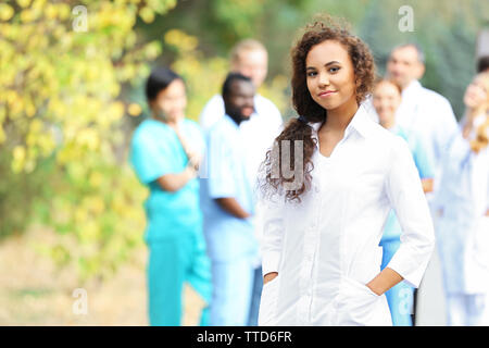 Attraente giovane donna medico con appunti in mani contro un gruppo di medici, all'aperto Foto Stock