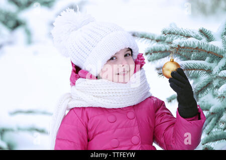 Bambina con abbigliamento invernale toccando golden ninnolo su abete nel parco innevato per esterno Foto Stock