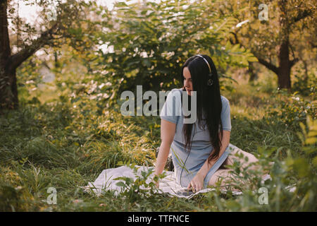 Premurosa donna con capelli lunghi ascoltare musica mentre è seduto sul campo erboso in giardino Foto Stock