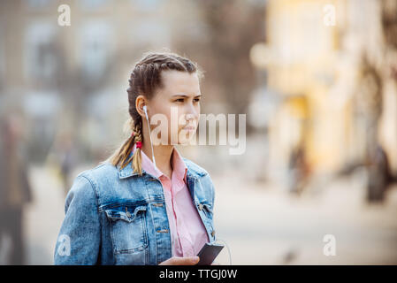 Ragazza in città ascoltando la musica dal telefono tramite le cuffie Foto Stock