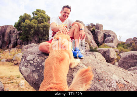 Sorridente metà uomo adulto petting cane mentre è seduto sulla roccia Foto Stock