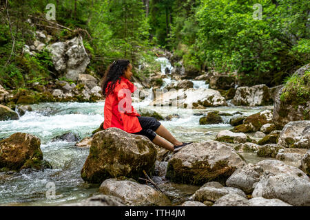 Giovane donna seduta su una roccia in un turchese fiume nella foresta Foto Stock