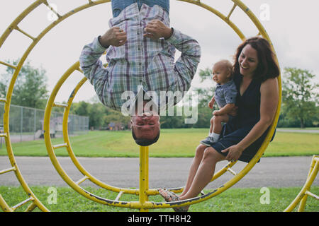 Madre e figlio guardando padre appeso circolare su Monkey bar nel parco giochi Foto Stock