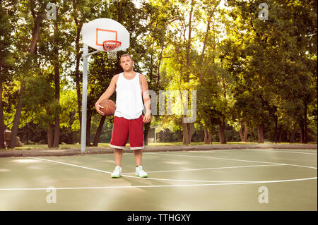 Ritratto di uomo con la pallacanestro in tribunale contro gli alberi in posizione di parcheggio Foto Stock