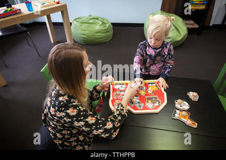 Insegnante assistere ragazza a manopola di gioco puzzle a tavola in età prescolare Foto Stock Insegnante assistere ragazza a manopola di gioco puzzle a tavola in età prescolare Foto Stock