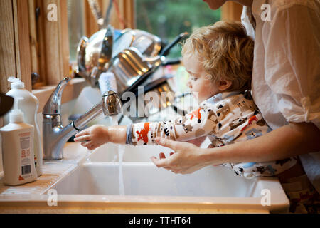 Immagine ritagliata della madre che assiste figlio di lavaggio delle mani al lavello da cucina Foto Stock