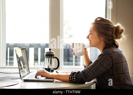 Donna felice utilizzando computer portatile mentre si gusta un caffè a tabella Foto Stock