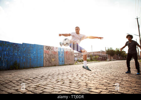 L'uomo calci palla calcio mentre amico guardando street Foto Stock