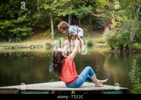 Donna felice figlio di sollevamento mentre è seduto sul molo al lago Foto Stock