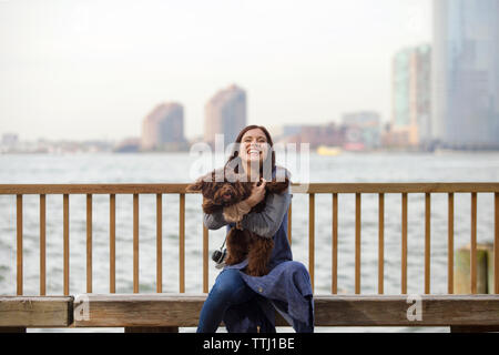 Giocoso donna con cane seduto sulla panchina contro il fiume Hudson Foto Stock