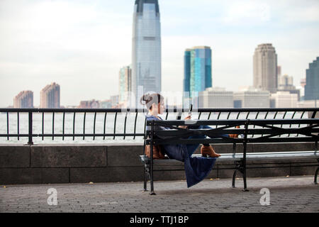 Vista laterale della donna che utilizza computer tavoletta mentre è seduto sulla panchina contro il fiume Hudson Foto Stock