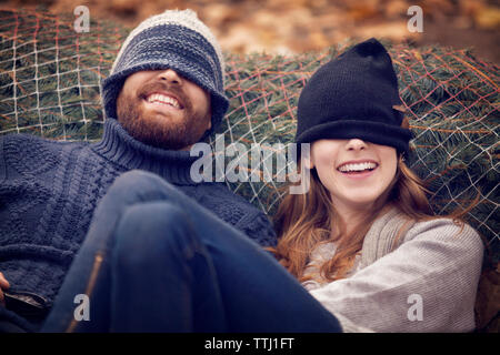 Coppia giocosa che indossa un cappello in maglia mentre si appoggia all'albero di natale in rete Foto Stock
