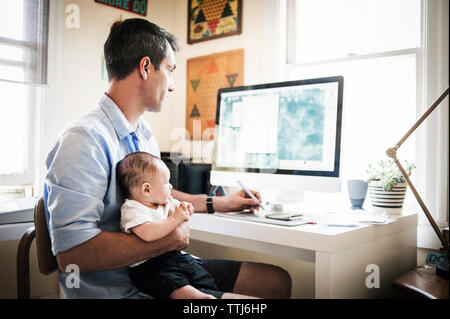 Uomo che porta bambino mentre si lavora su un computer desktop a casa Foto Stock