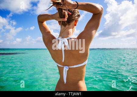 Vista posteriore della donna in bikini top regolazione capelli mentre in piedi in mare contro il cielo nuvoloso Foto Stock