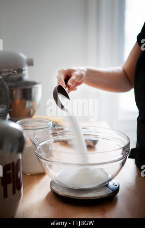 Immagine ritagliata della donna versando la farina in un recipiente mantenuto sulla bilancia da cucina Foto Stock