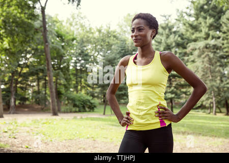 Donna sorridente con le mani sul hip guardando lontano mentre si sta in piedi sul campo nel parco Foto Stock