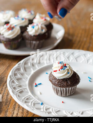 Il cioccolato mini cupcake sulla piastra bianca decorata con rosso, bianco e blu spruzza. Dita con motion blurr in foto. Decorate aggiuntivi Foto Stock