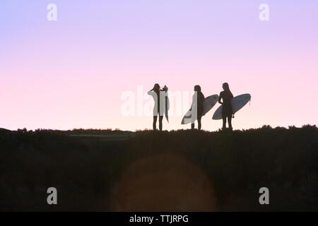 Silhouette amiche tenendo la tavola da surf mentre si sta in piedi sul campo durante il tramonto Foto Stock