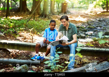 Insegnante che mostra libro studente mentre è seduto su passerella in foresta Foto Stock