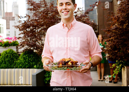 Sezione mediana di uomo sorridente che trasportano il pollo arrosto nel vassoio mentre in piedi contro piante Foto Stock