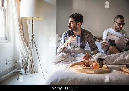 Gay man aventi la prima colazione mentre il partner la lettura di giornale sul letto di casa Foto Stock