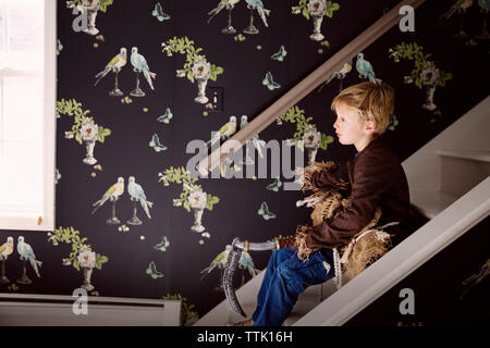 Vista laterale del ragazzo costume di contenimento mentre è seduto sulla scalinata a casa Foto Stock