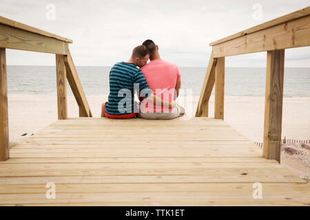 Vista posteriore della coppia omosessuale seduti sul Boardwalk contro il mare Foto Stock