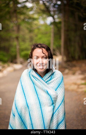 Ritratto di donna sorridente avvolto in una coperta permanente sulla strada in foresta Foto Stock