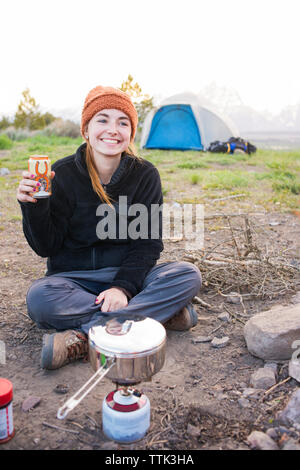 Donna sorridente holding drink seduto al campeggio Foto Stock