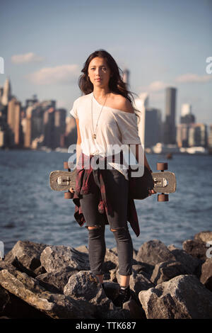 Ritratto di donna azienda skateboard sulla spiaggia rocciosa contro sky Foto Stock