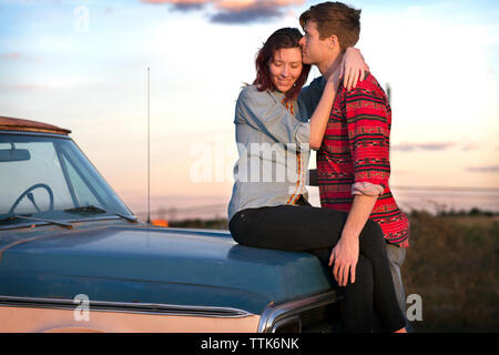 Ragazza abbracciando il ragazzo seduto sul cofano del veicolo contro sky Foto Stock