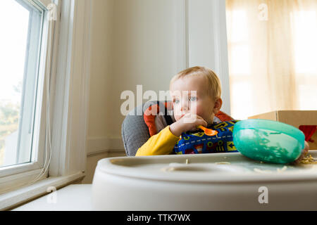 Carino premurose baby boy con recipiente di plastica e versare seduto sulla sedia alta a casa Foto Stock