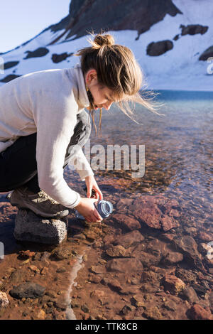 Elevato angolo di visione femminile di escursionista il riempimento di acqua in bottiglia dal fiume Foto Stock