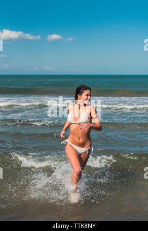 Donna felice indossando bikini bianco mentre è in esecuzione la mare contro il cielo blu durante la giornata di sole Foto Stock