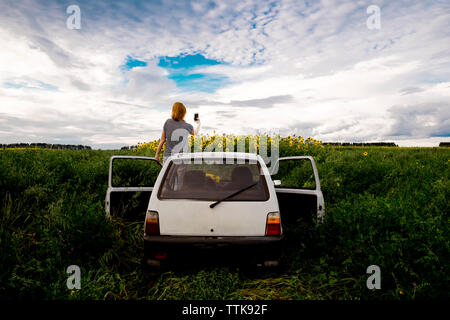 Vista posteriore della donna di fotografare i girasoli sul campo mentre in piedi in auto contro il cielo nuvoloso Foto Stock