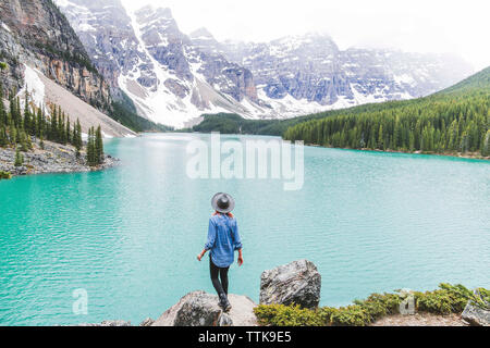 Vista posteriore della donna in fedora hat permanente sulla roccia tramite il Lago Moraine contro le montagne Foto Stock