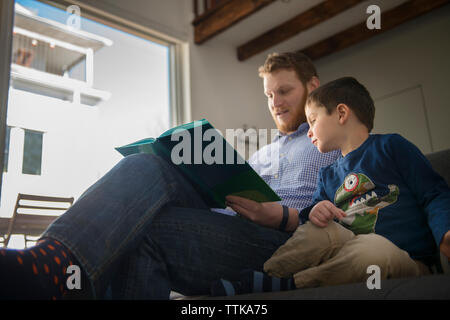 Basso angolo di visione del padre azienda prenota e figlio di insegnamento sul divano Foto Stock