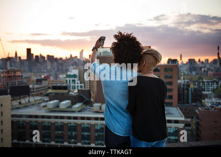 Vista posteriore del giovane tenendo selfie mentre si sta in piedi sul tetto durante il tramonto Foto Stock