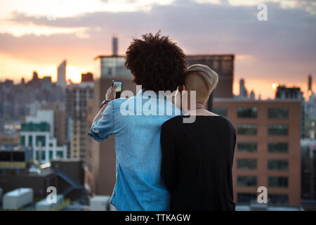 Vista posteriore del giovane in piedi con telefono cellulare sul tetto durante il tramonto Foto Stock