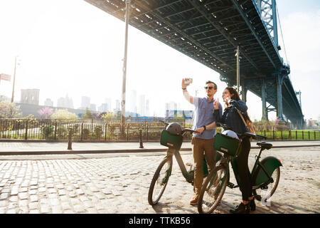 Giovane tenendo selfie stando in piedi con le biciclette contro Manhattan bridge Foto Stock