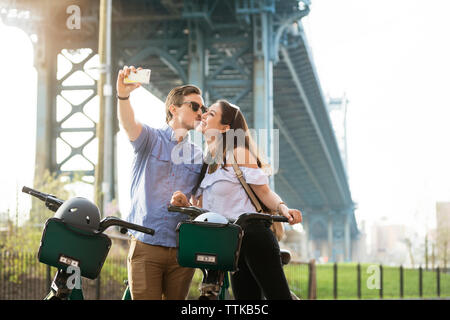 Giovane kissing tenendo selfie con biciclette contro Manhattan Bridge Foto Stock