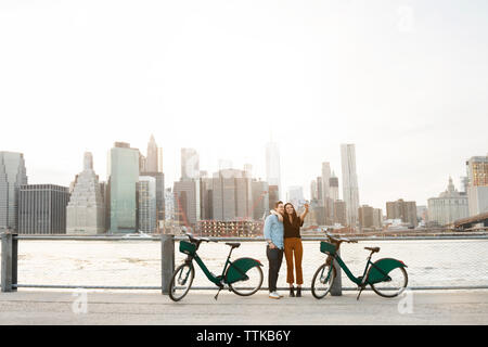 Giovane tenendo selfie permanente, mentre da parte delle biciclette contro East River in città Foto Stock