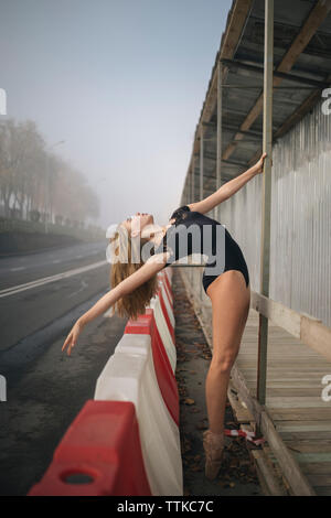 Vista laterale della ballerina vestita di nero leotard stretching al marciapiede in città Foto Stock