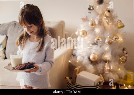 Ragazza felice che tiene biscotti e bicchiere di latte mentre sta accanto all'albero di natale a casa Foto Stock