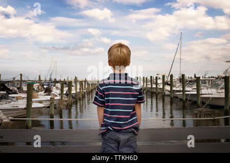 Vista posteriore del ragazzo in piedi sul ponte mediante harbour contro il cielo nuvoloso Foto Stock