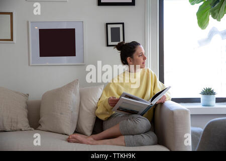 Donna che mantiene magazine che guarda lontano mentre è seduto sul divano di casa Foto Stock